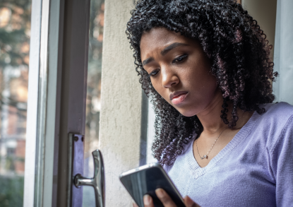 Eine junge Schwarze Frau steht vor einem Fenster und schaut traurig auf ihr Smartphone.